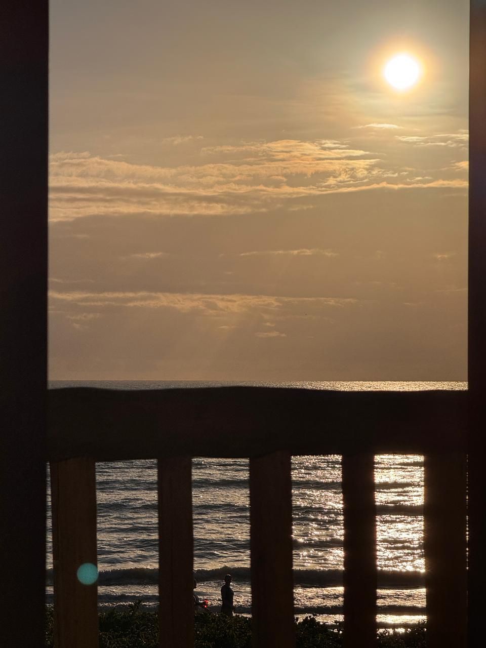 A silhueta de uma pessoa se destaca na praia ao nascer do sol, vista através da grade de uma varanda.
