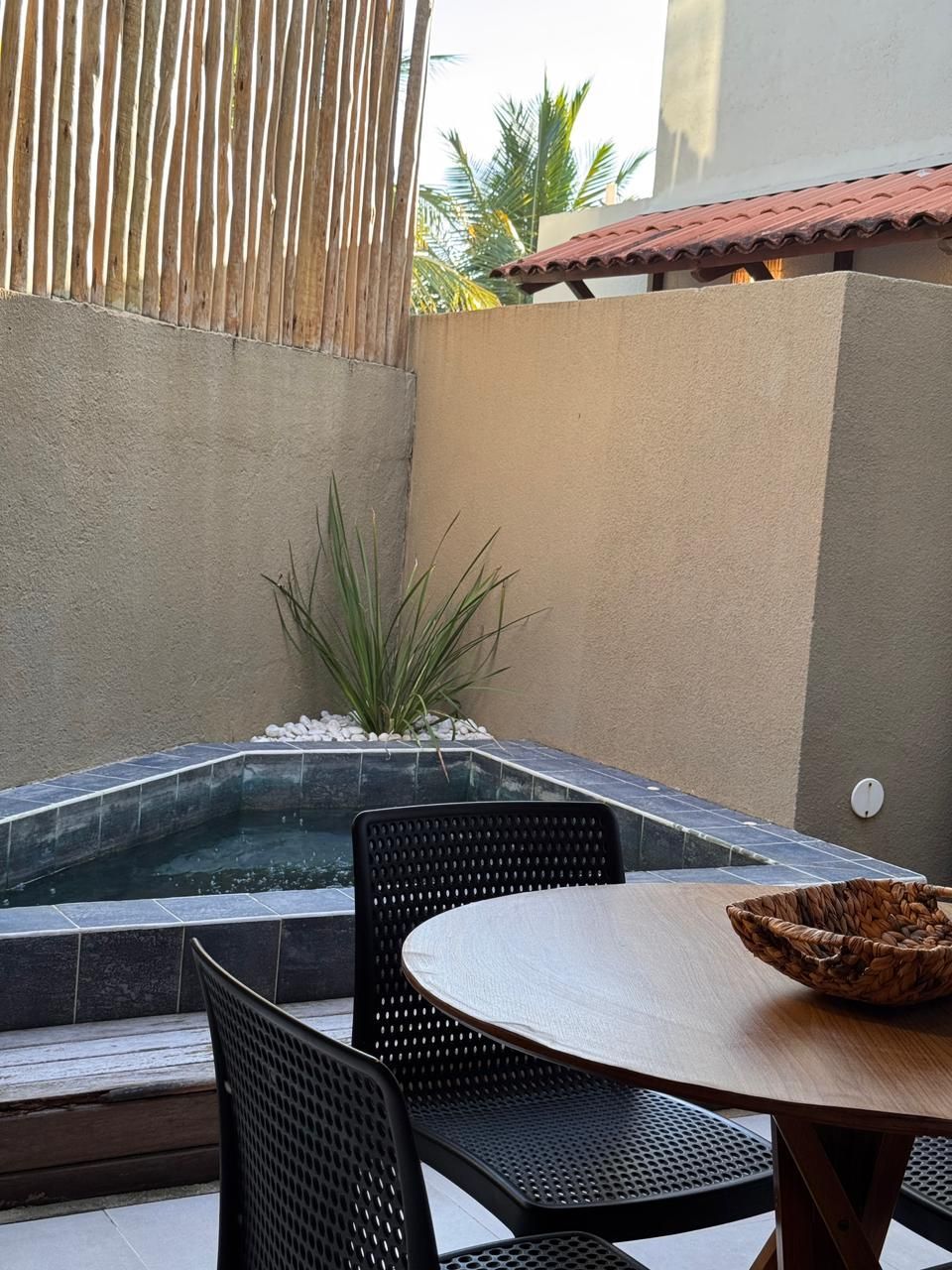 Uma pequena piscina rasa com uma planta, ao lado de uma mesa redonda de madeira e cadeiras pretas em um pátio com divisória de bambu.