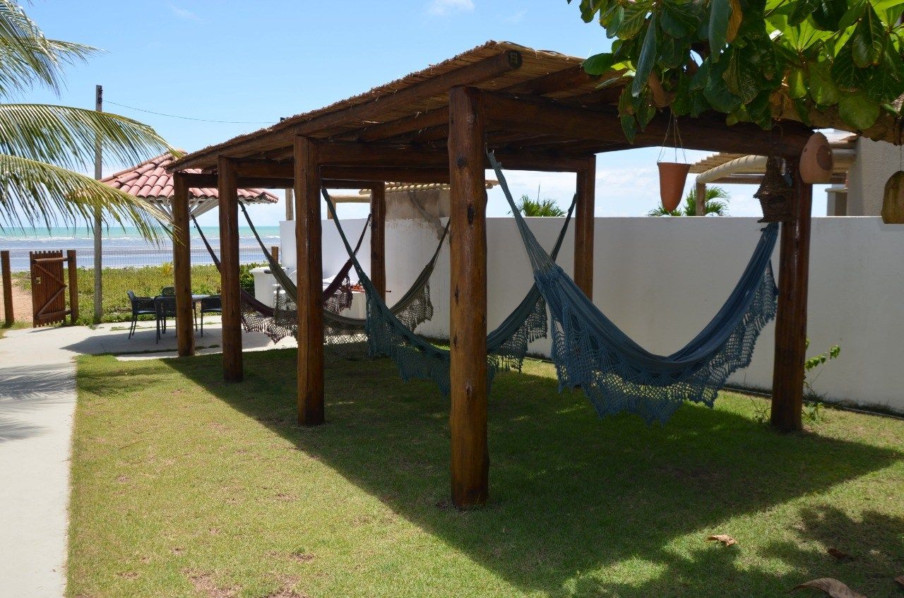 Uma pérgola de madeira ao ar livre apresenta várias redes azuis suspensas sobre um gramado à beira de uma praia costeira.