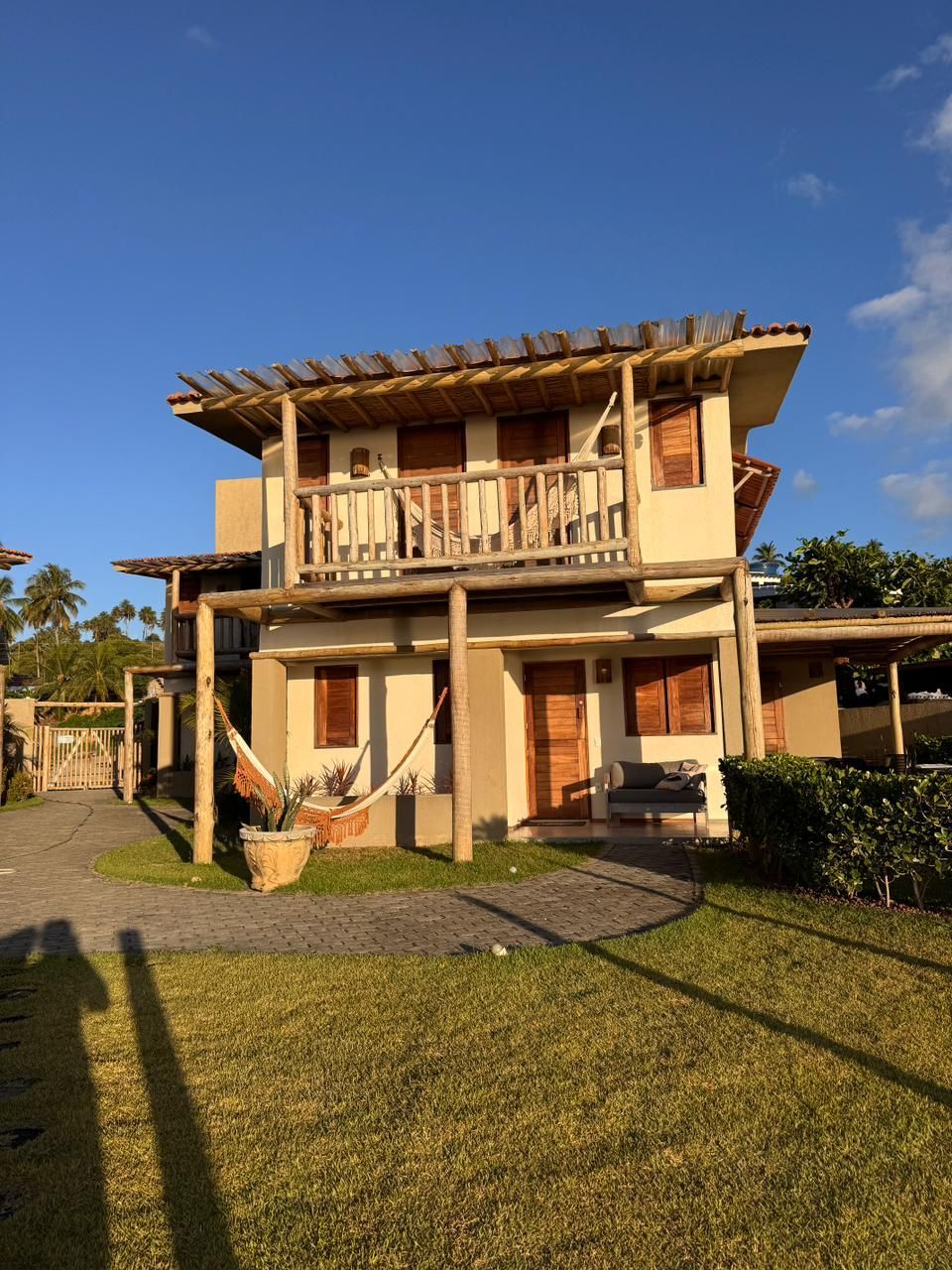 Casa costeira de dois andares em tons de bege, com varanda de madeira, área de estar externa e rede sob um céu azul límpido.