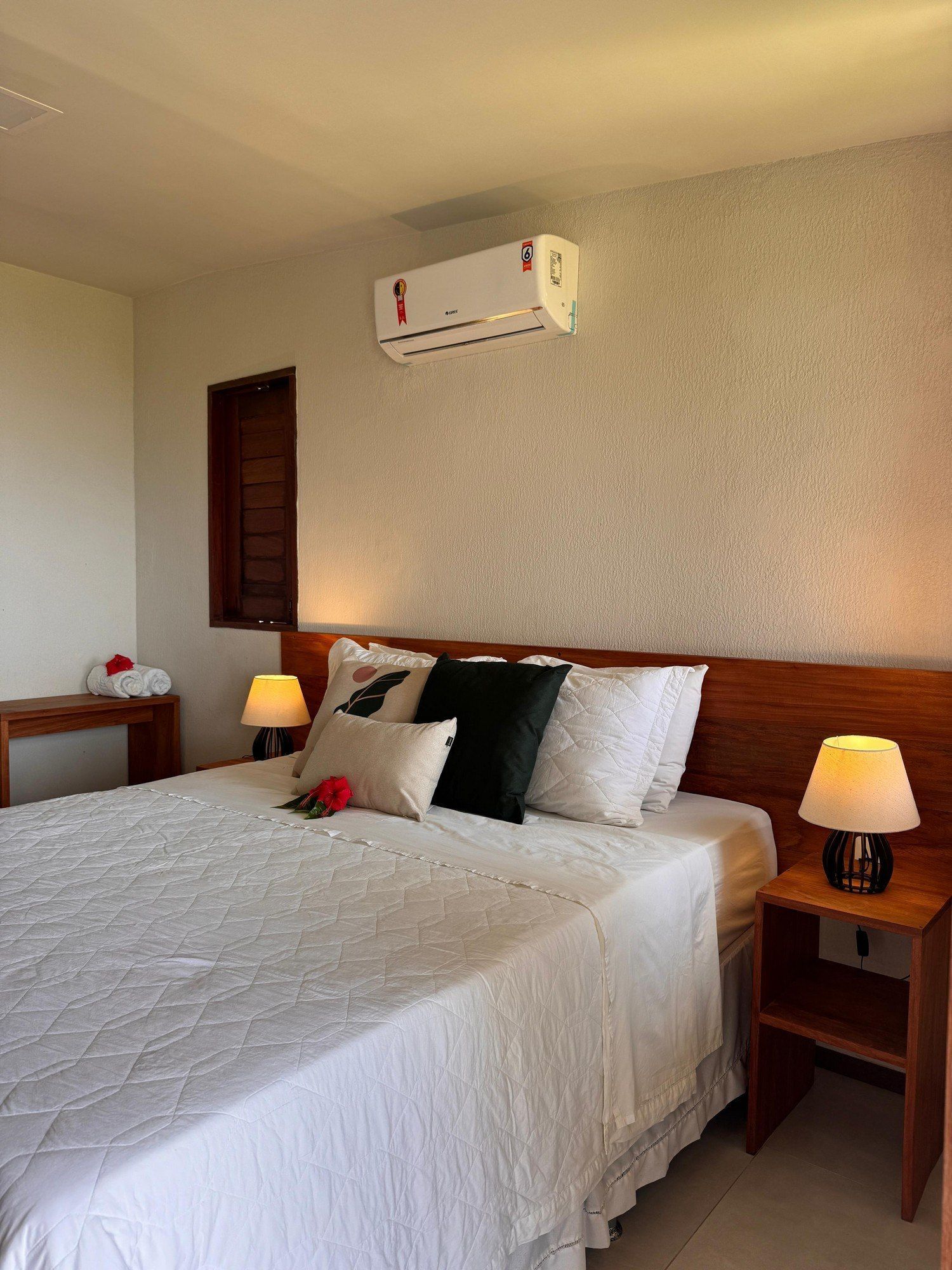 Um quarto com uma cama com roupa de cama branca, almofadas variadas, duas mesas de cabeceira de madeira com abajures e um ar-condicionado de parede.
