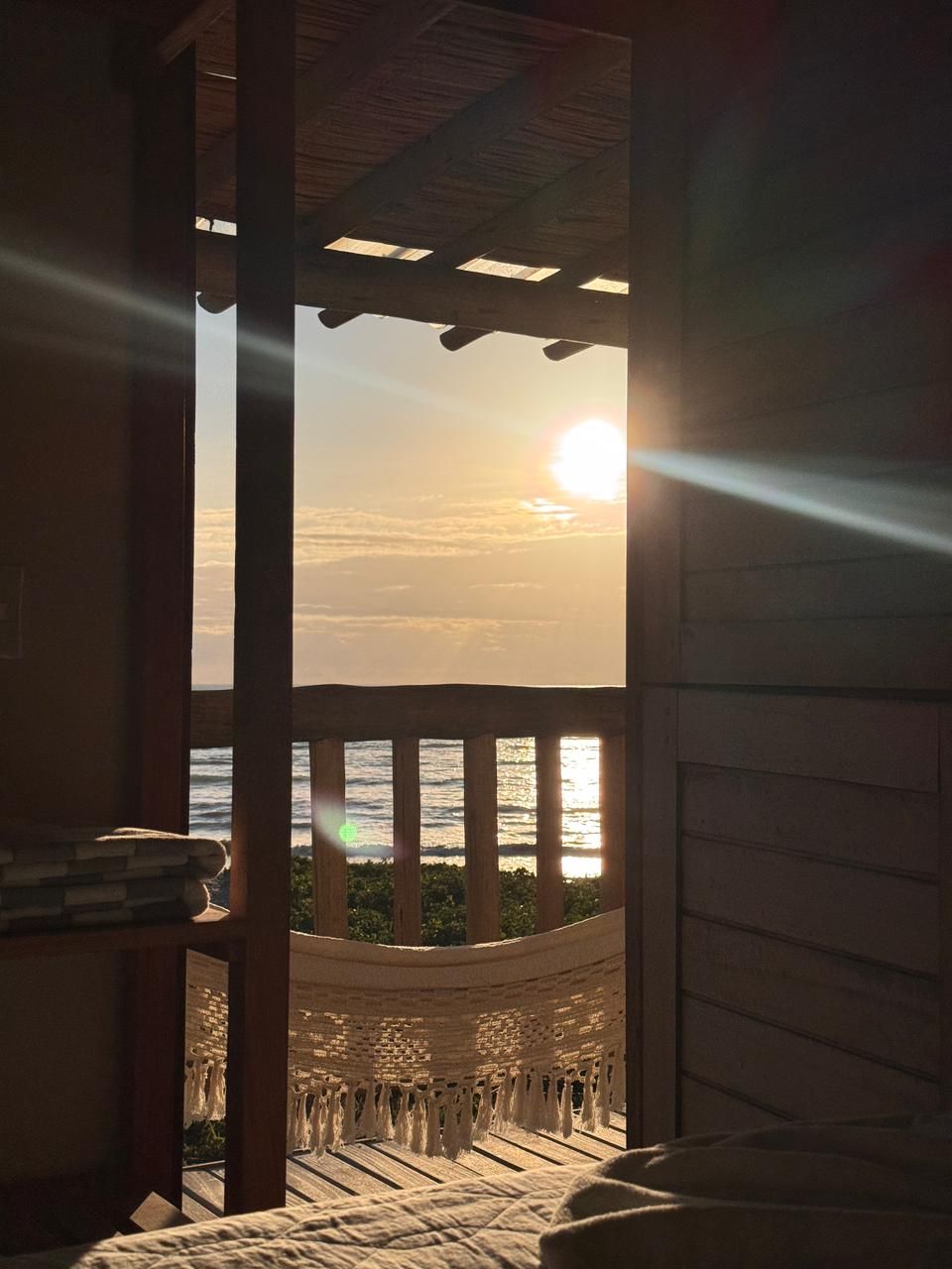 Vista ensolarada da varanda para o oceano ao nascer do sol, com uma rede suspensa e um deck de madeira, vista de dentro de um quarto.