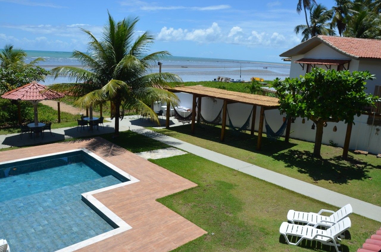 Uma piscina num pátio ao lado de um gramado com uma pérgola com rede e duas espreguiçadeiras, com vista para a praia e o oceano.