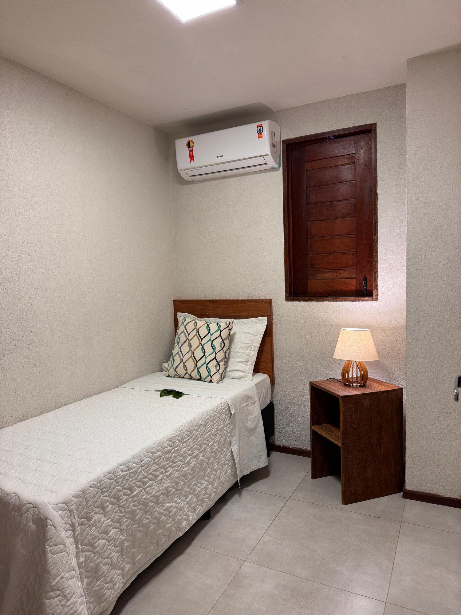 Um pequeno quarto com uma cama de solteiro com uma almofada estampada, uma mesa de cabeceira de madeira com um abajur e um aparelho de ar condicionado fixado na parede.