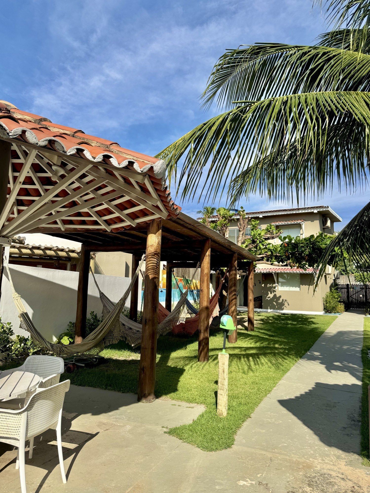 Uma pérgola de madeira com várias redes fica em um gramado ao lado de uma passarela pavimentada e um prédio de dois andares sob um céu azul.