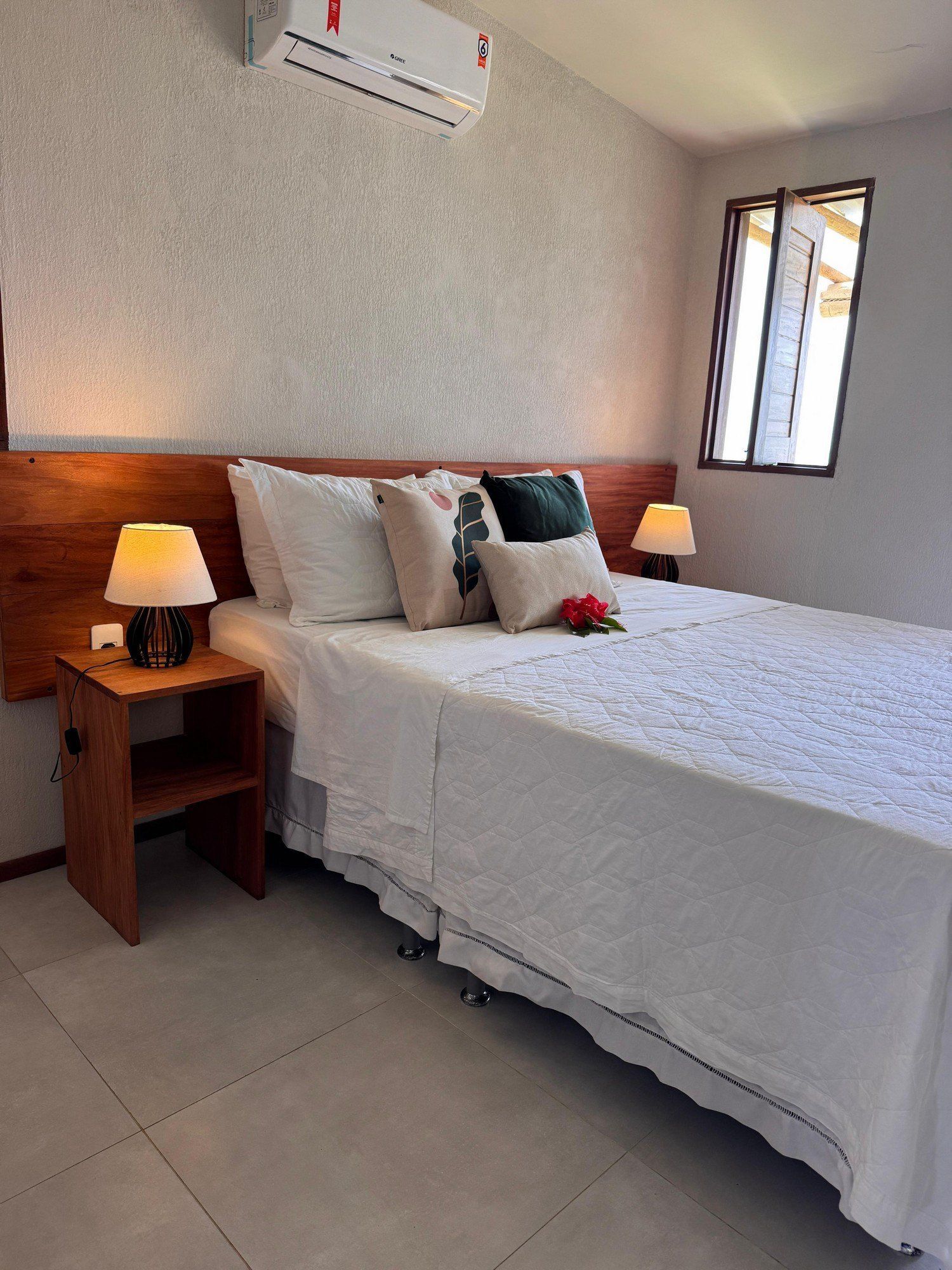 Um quarto impecável com cama de lençóis brancos, vários travesseiros, cabeceira de madeira, mesa de cabeceira com abajur e ar-condicionado.
