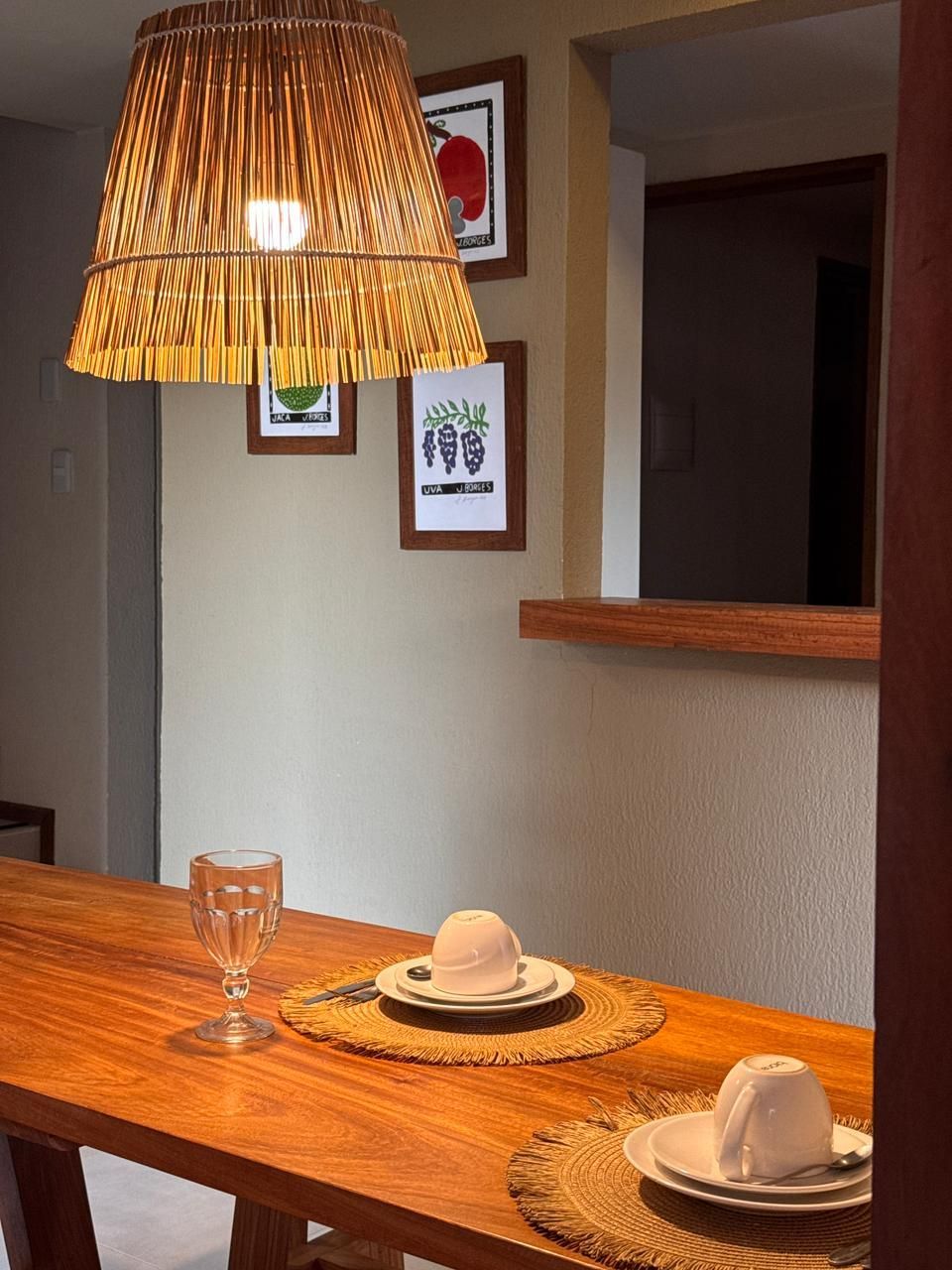 Uma mesa de jantar de madeira com dois lugares, um candeeiro de teto trançado e obras de arte emolduradas na parede.