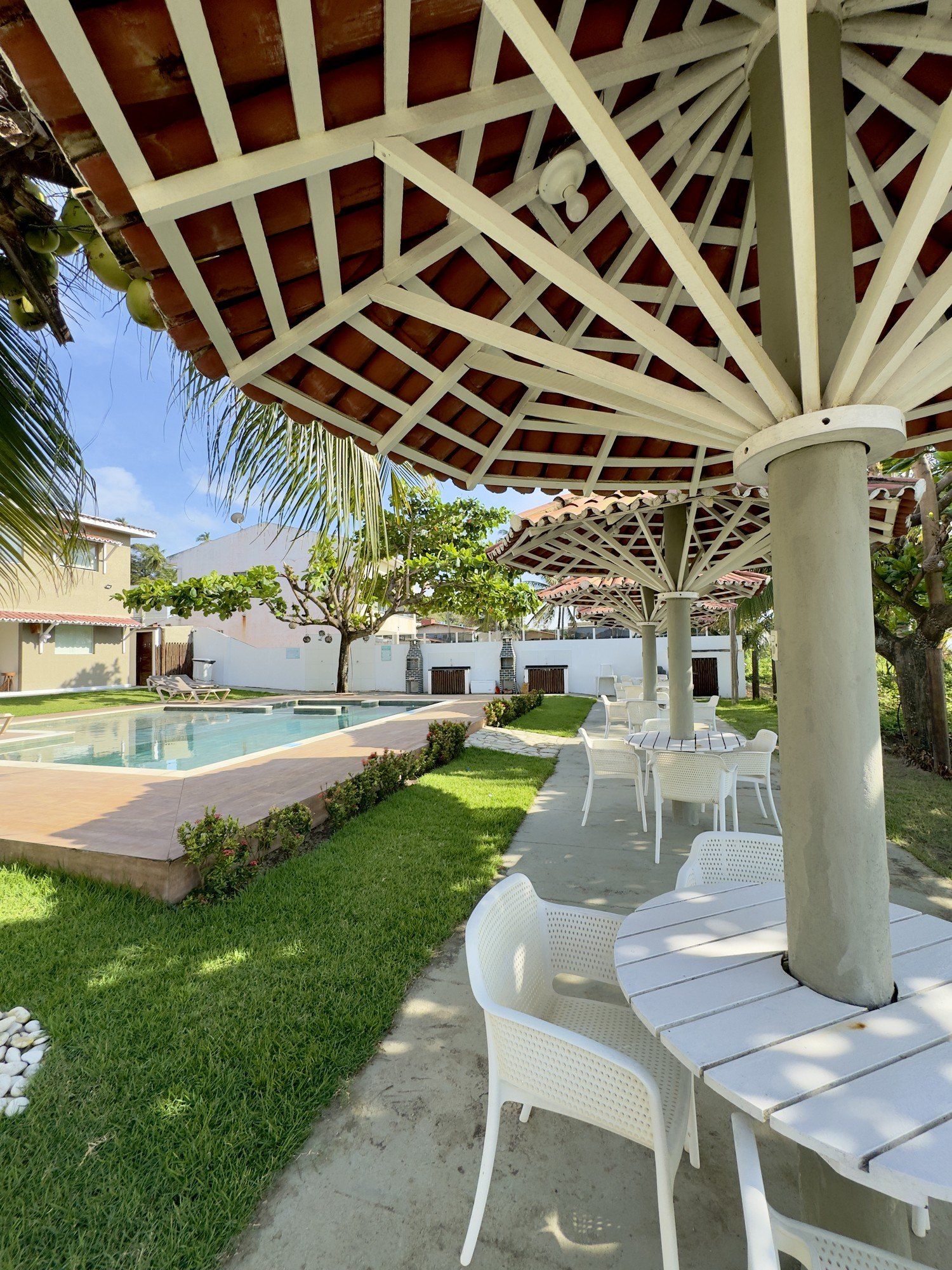 Mesas e cadeiras brancas sob grandes guarda-sóis junto a uma piscina exterior num dia ensolarado.