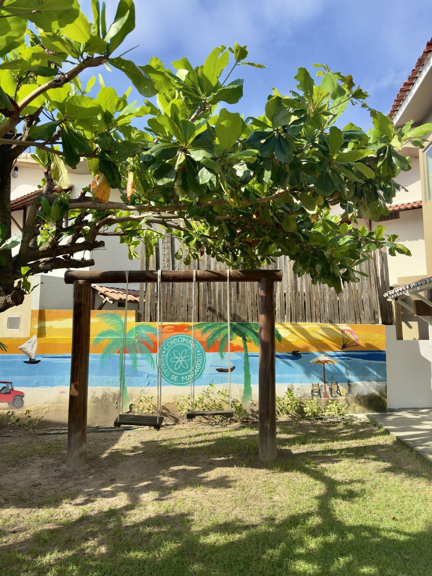 Um balanço de madeira está instalado em um gramado sob uma grande árvore, em frente a um mural que retrata uma ensolarada cena de praia.