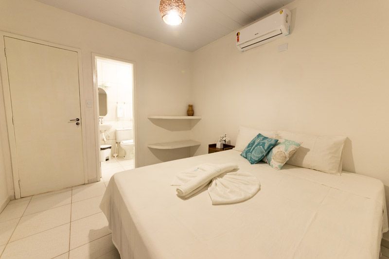 Um quarto de hotel com uma cama de lençóis brancos, duas pequenas prateleiras suspensas e uma porta aberta para o banheiro.