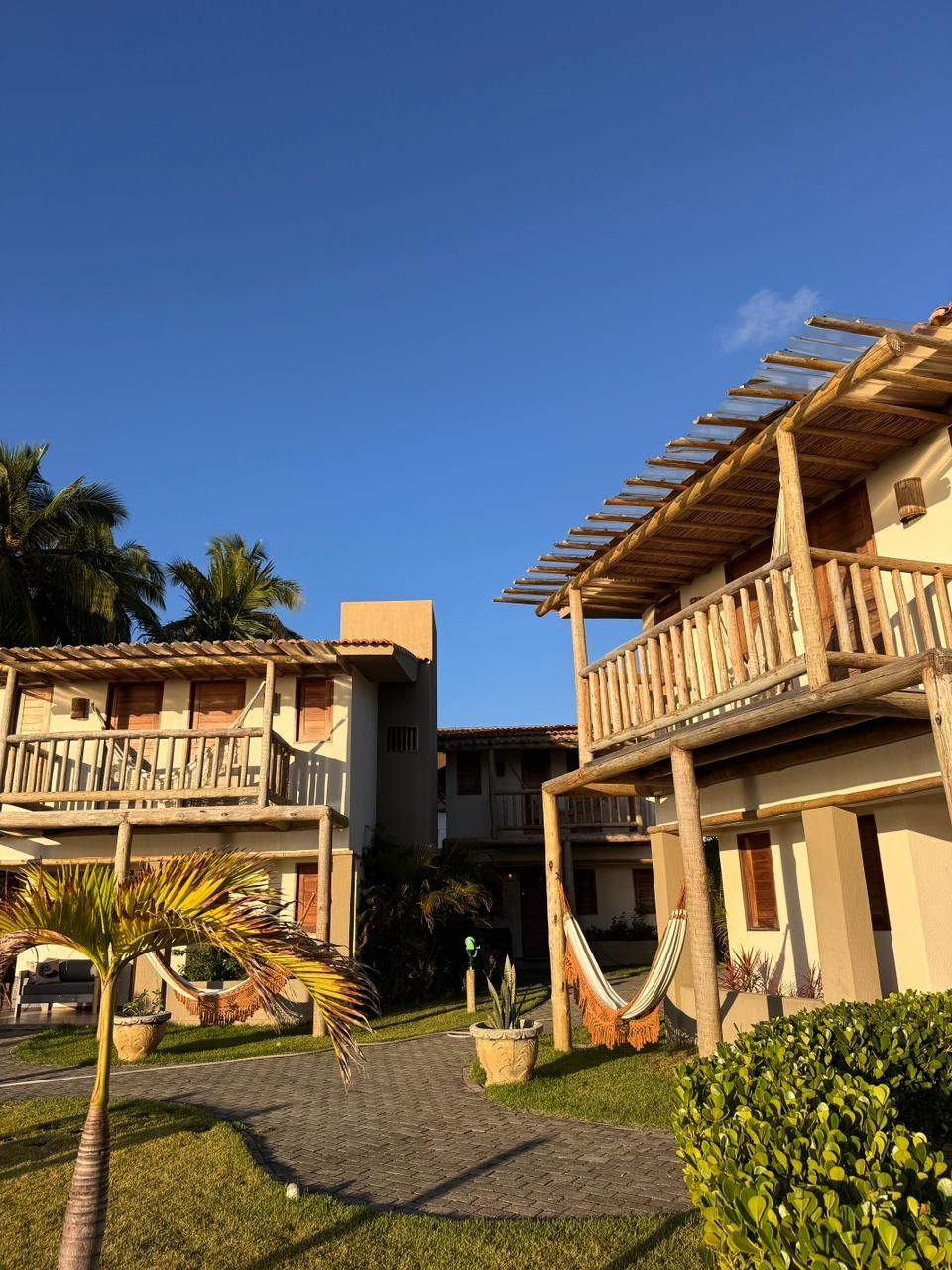 Casas de madeira de dois andares para férias, com varandas e redes, dispostas em torno de um pátio gramado sob um céu azul límpido.
