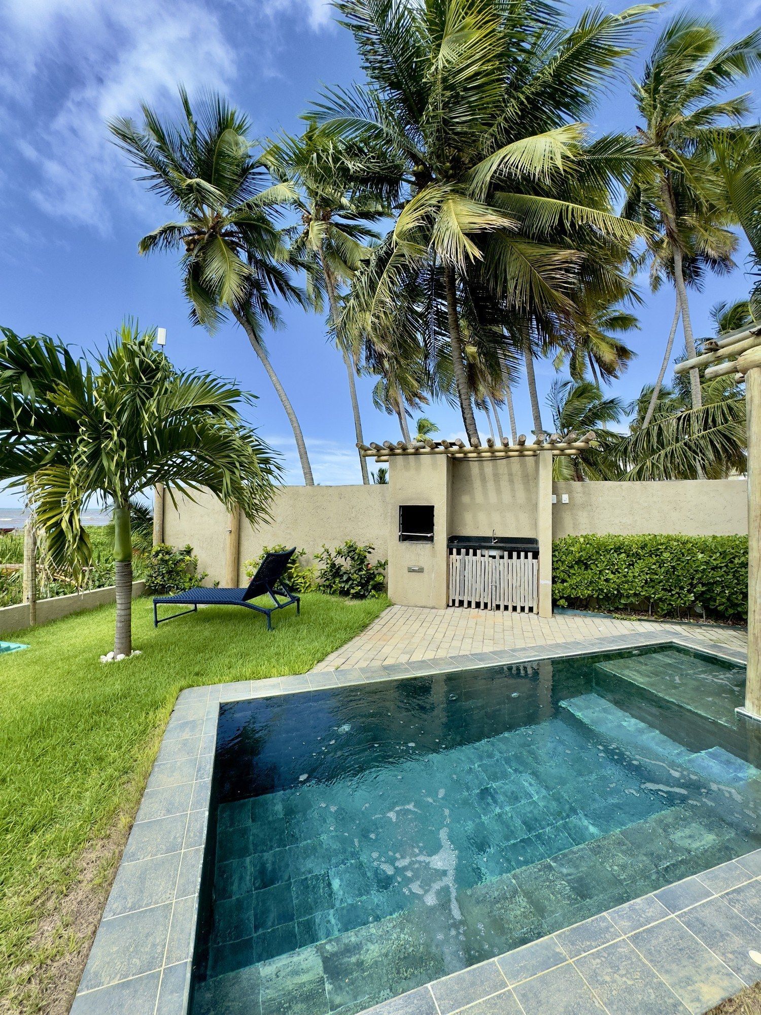 Uma pequena piscina revestida com azulejos escuros fica ao lado de um pátio, uma espreguiçadeira e palmeiras sob um céu azul brilhante.