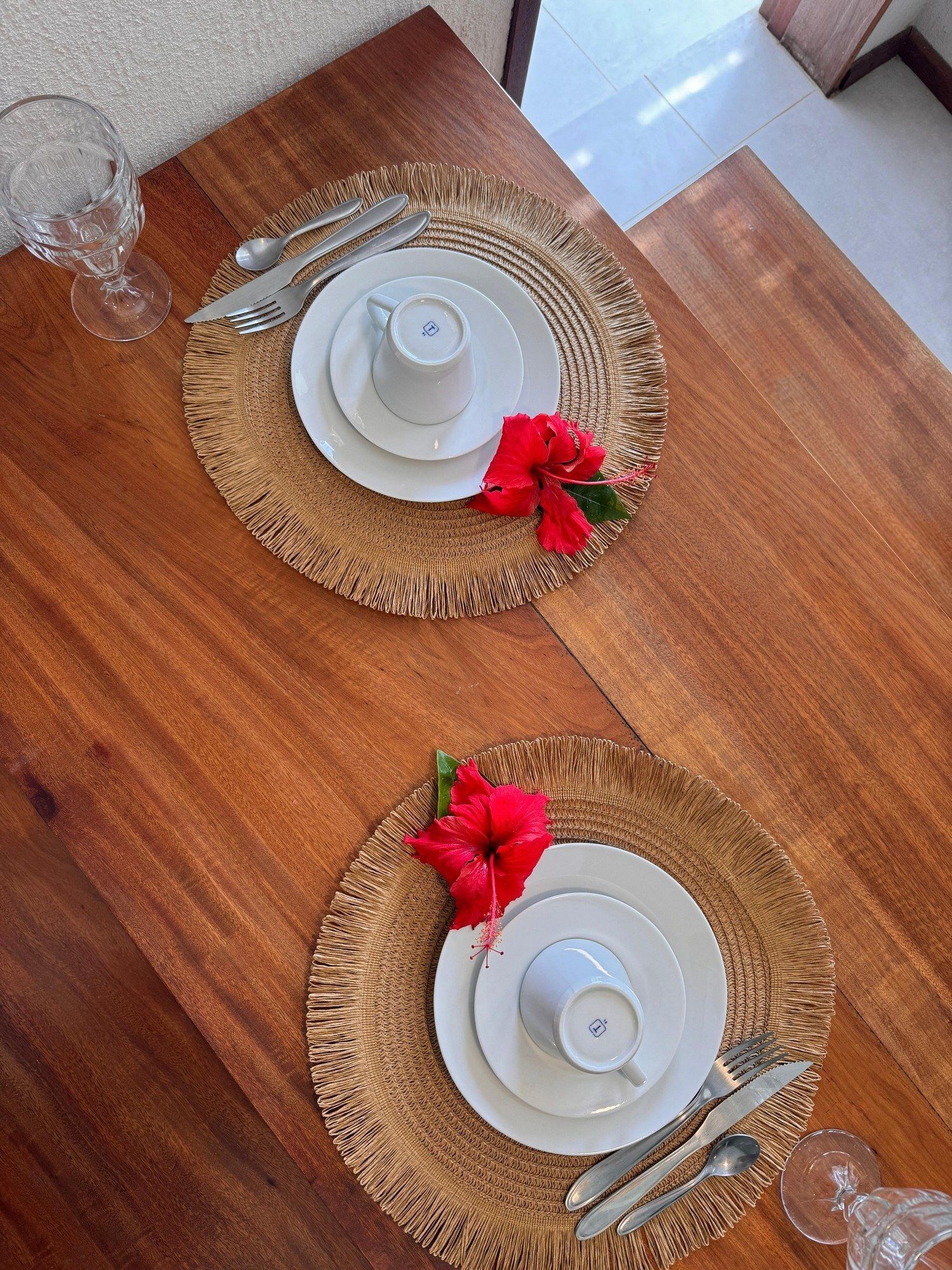 Dois lugares à mesa com pratos brancos, flores de hibisco vermelhas e talheres sobre jogos americanos redondos de vime, em uma mesa de madeira.
