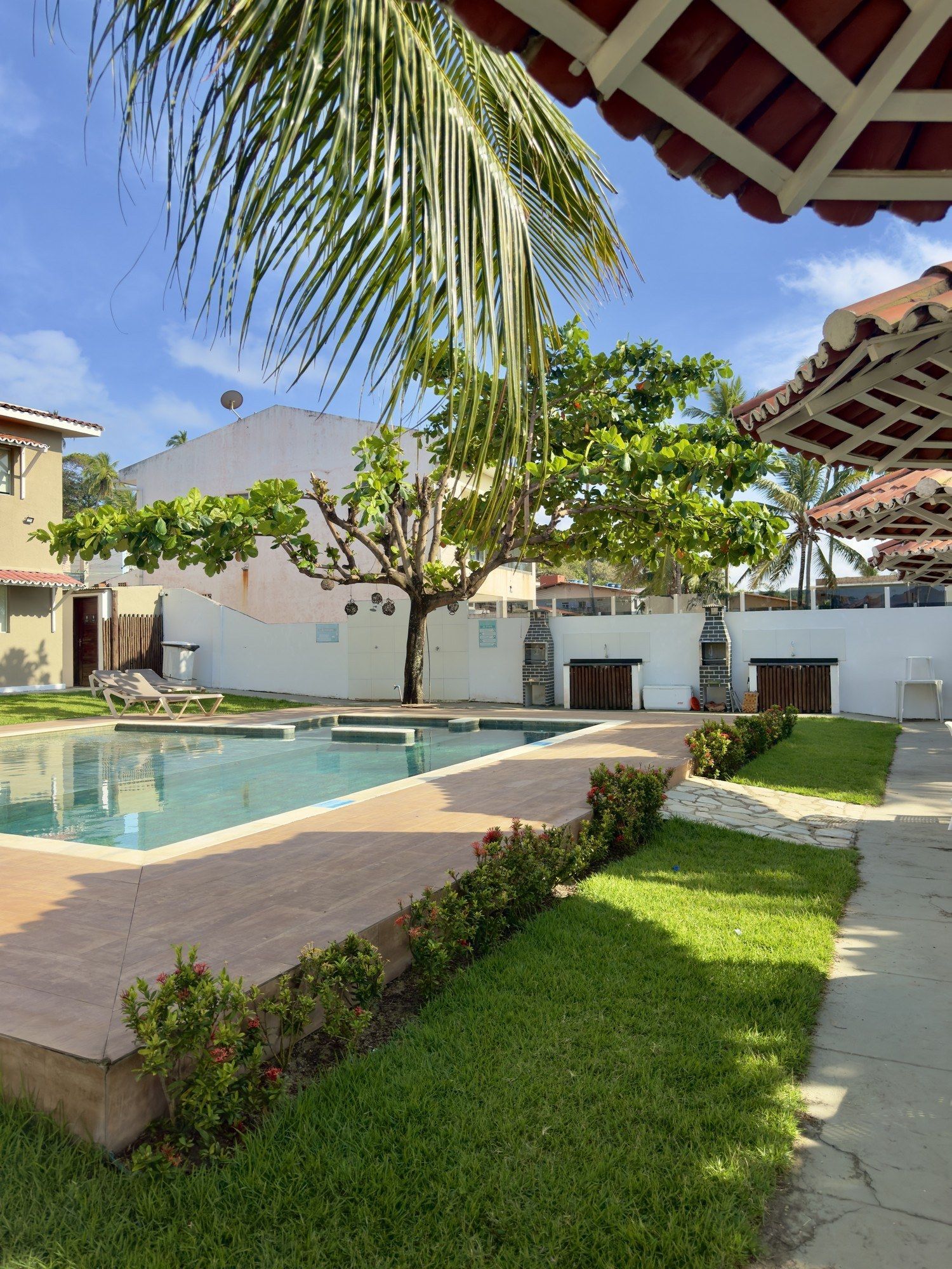 Uma piscina externa iluminada, rodeada por grama verde, um deck de azulejos e uma grande árvore sob um céu azul claro.