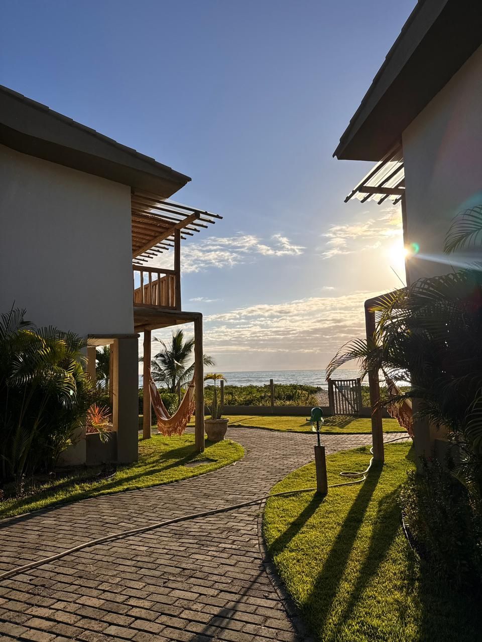 Um caminho pavimentado conduz entre dois edifícios costeiros em direção ao oceano ao nascer do sol, com redes visíveis nas varandas.