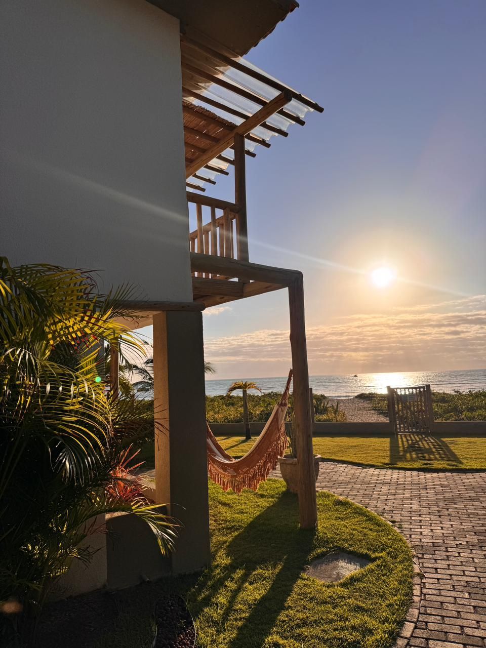 Uma casa de férias tropical com varanda e rede oferece vista para uma praia oceânica ensolarada durante o pôr do sol.