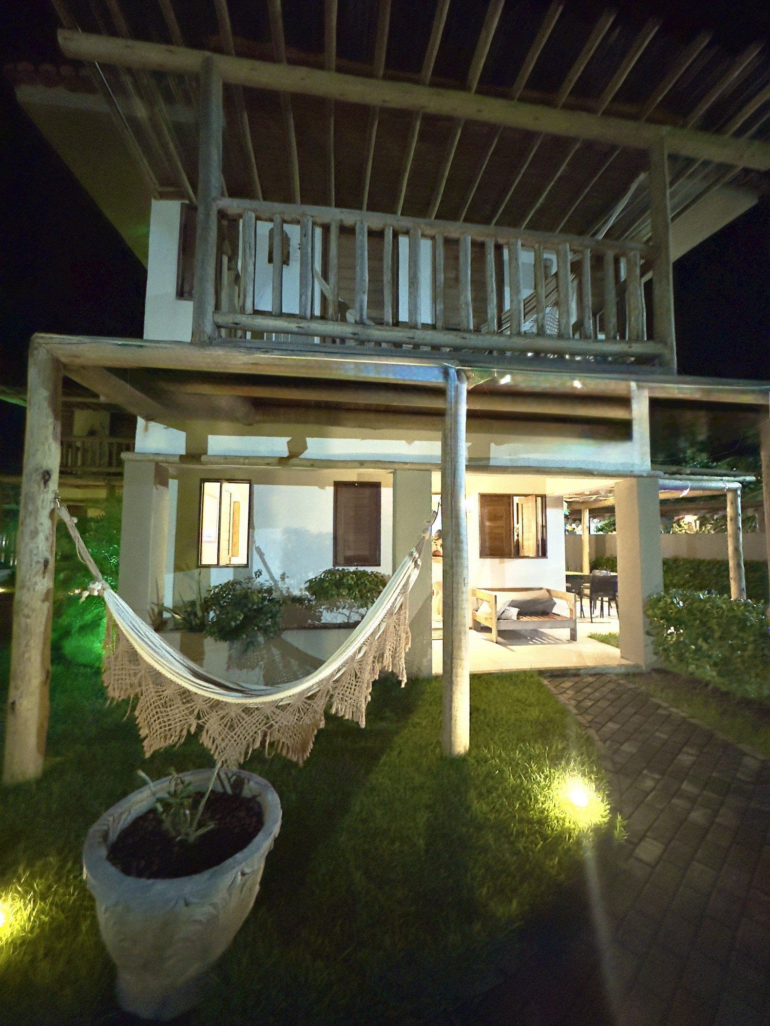 Uma casa rústica de dois andares à noite, com uma rede em primeiro plano, o térreo iluminado e uma varanda de madeira no andar superior.