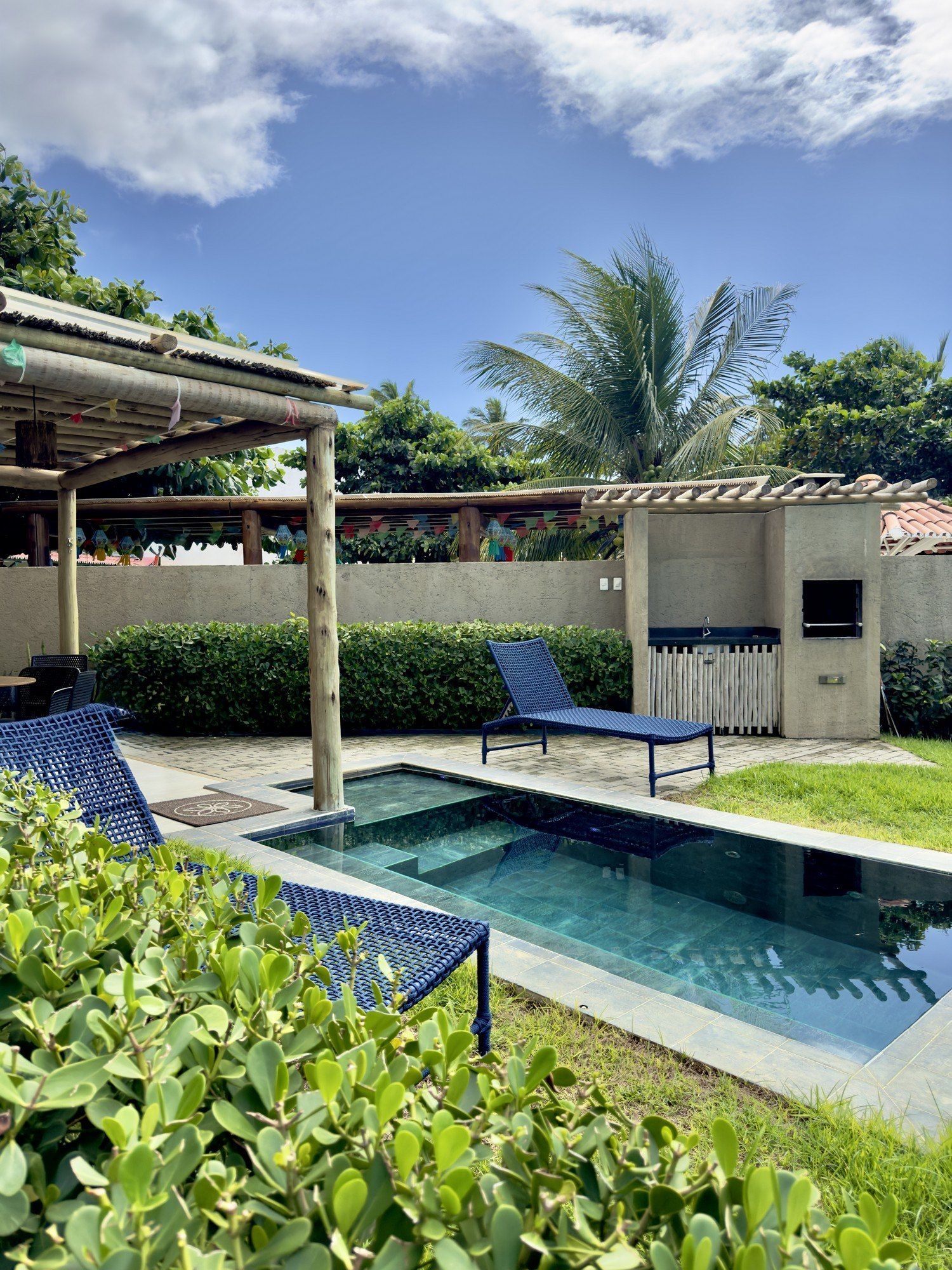 Uma piscina retangular no quintal, duas espreguiçadeiras azuis e uma pequena área de churrasqueira ao ar livre sob um céu azul e ensolarado.