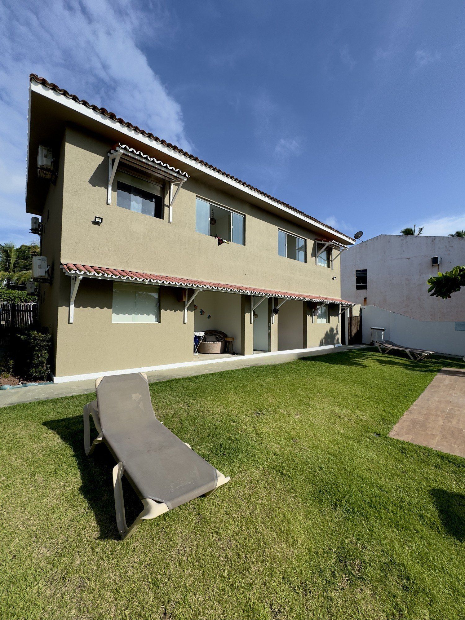 Um prédio de apartamentos de dois andares, cor bege, com um gramado e uma solitária espreguiçadeira sob um céu azul brilhante.