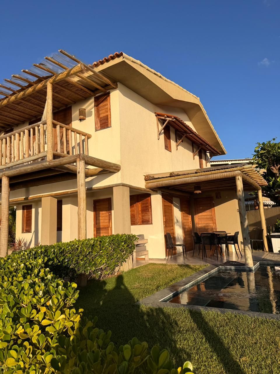 Uma casa de praia de dois andares, com fachada de estuque, varanda de madeira e pérgola, rodeada de vegetação e uma pequena piscina sob um céu azul.