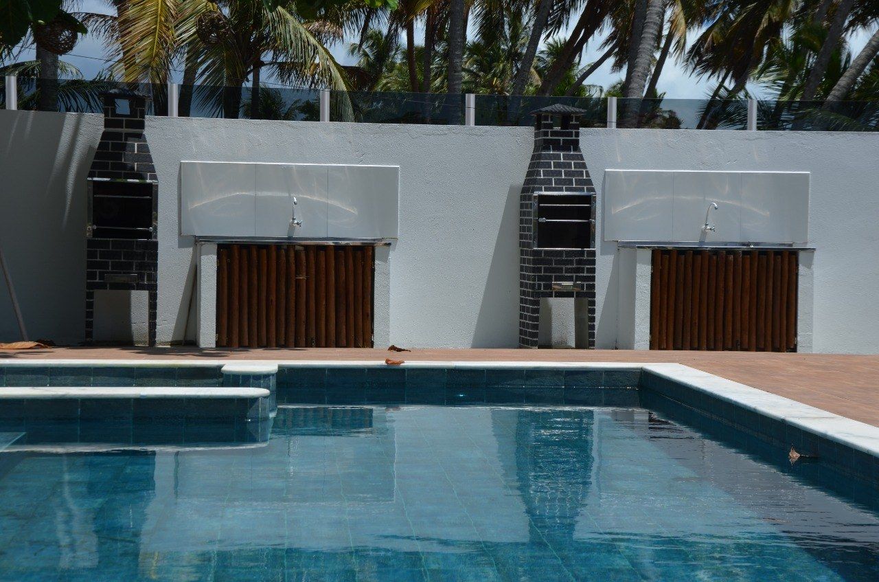 Duas churrasqueiras externas de tijolos e bancadas de preparo em aço inoxidável ficam ao lado de uma piscina, encostadas em uma parede branca.