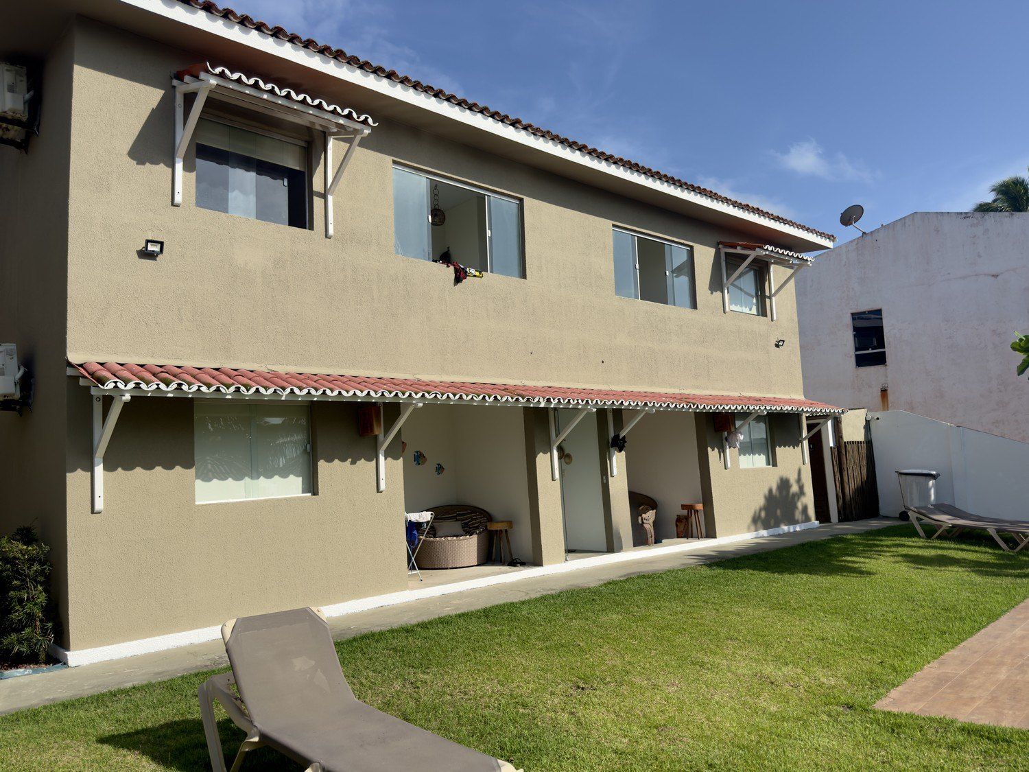 Um prédio de apartamentos de dois andares, cor bege, com um pátio coberto, toldo de telhas vermelhas e um gramado na frente.