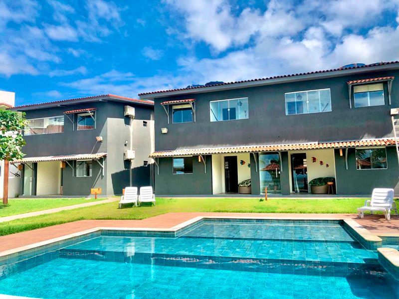 Duas modernas casas de férias em tons de cinza escuro têm vista para uma piscina azul brilhante em um dia ensolarado com céu parcialmente nublado.