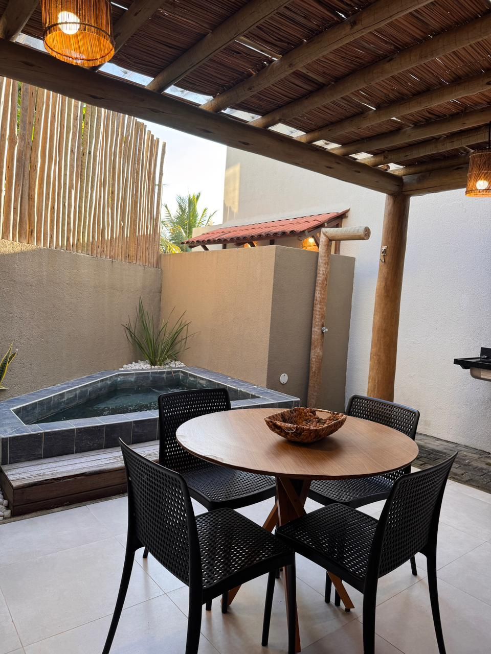 Um pátio com uma mesa redonda de madeira, quatro cadeiras pretas e uma pequena piscina embutida sob um telhado de ripas de bambu.