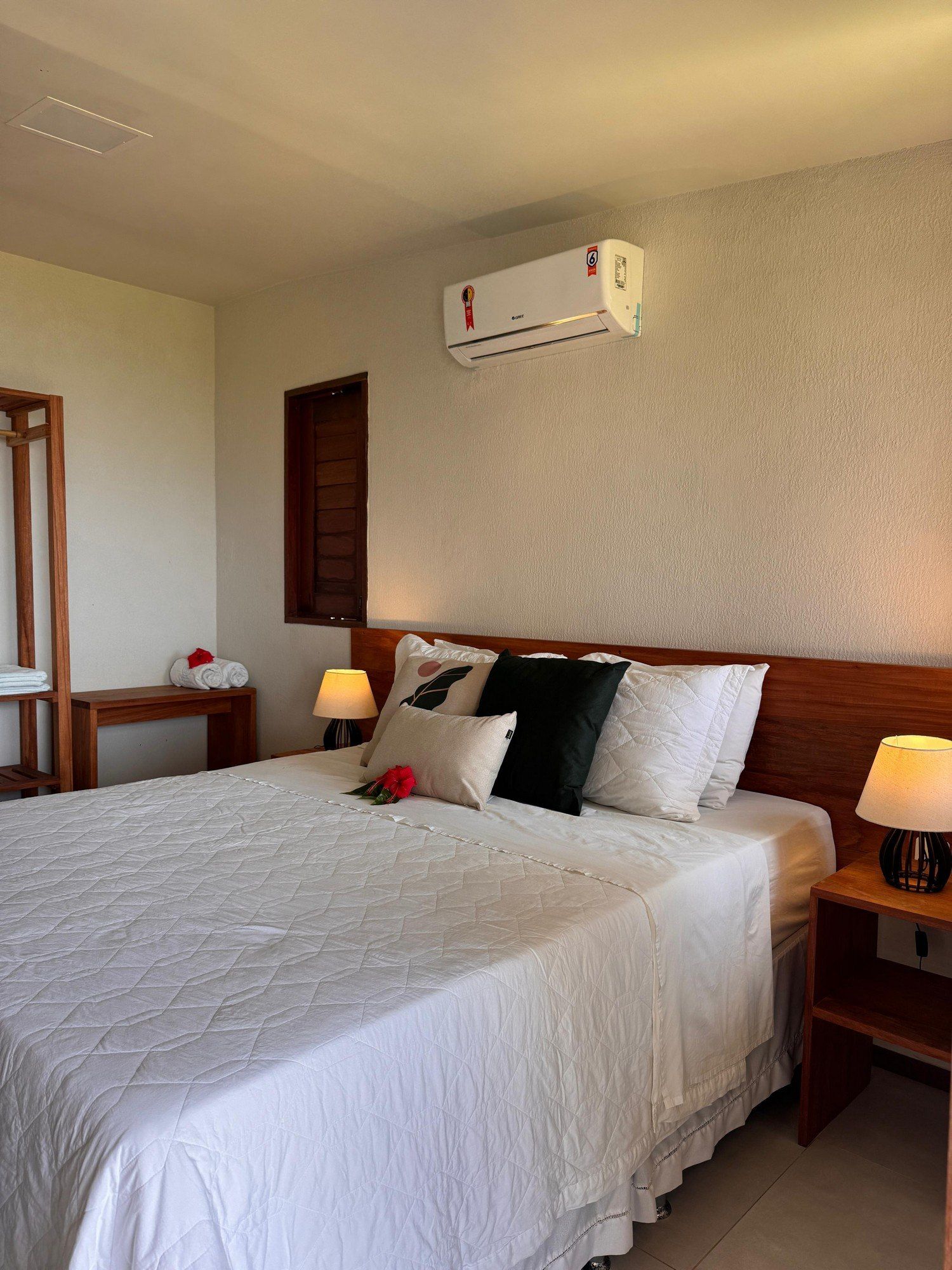 Um quarto de hotel com cama queen-size, roupa de cama branca, travesseiros pretos e brancos, móveis de madeira e ar-condicionado de parede.
