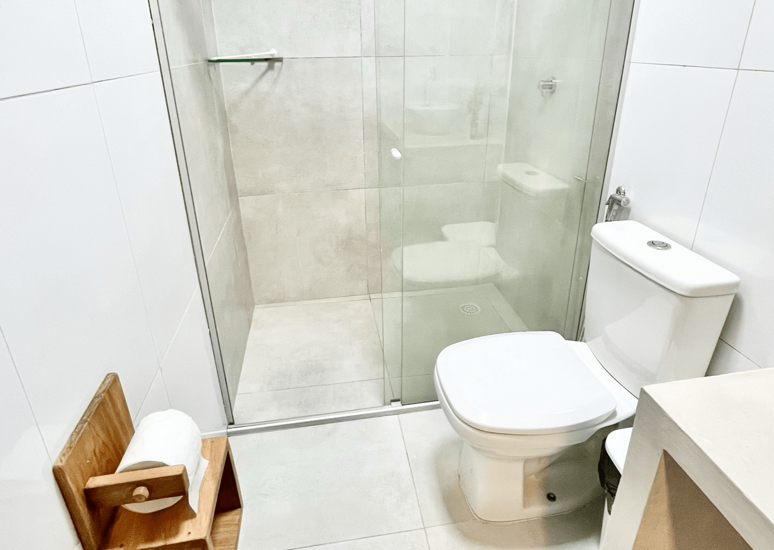 Um banheiro moderno com azulejos brancos, box de vidro para o chuveiro, vaso sanitário e um pequeno porta-papel higiênico de madeira.