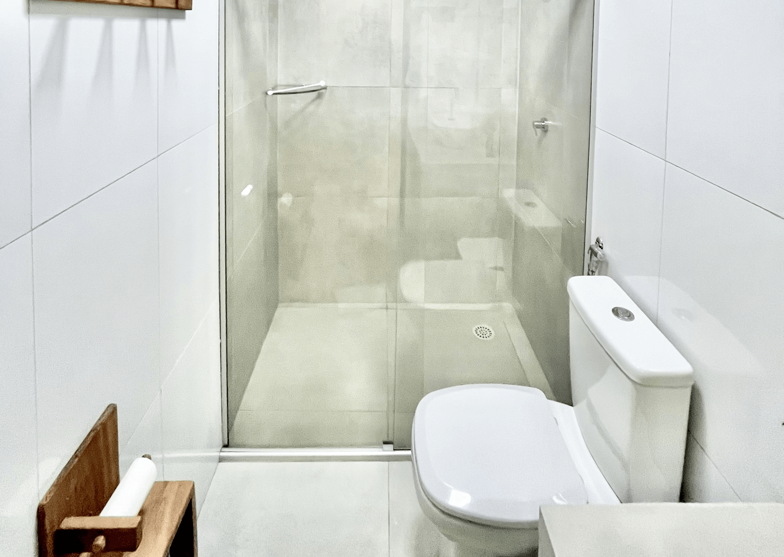 Um banheiro moderno e branco com box de vidro, vaso sanitário branco e um pequeno porta-papel higiênico de madeira.