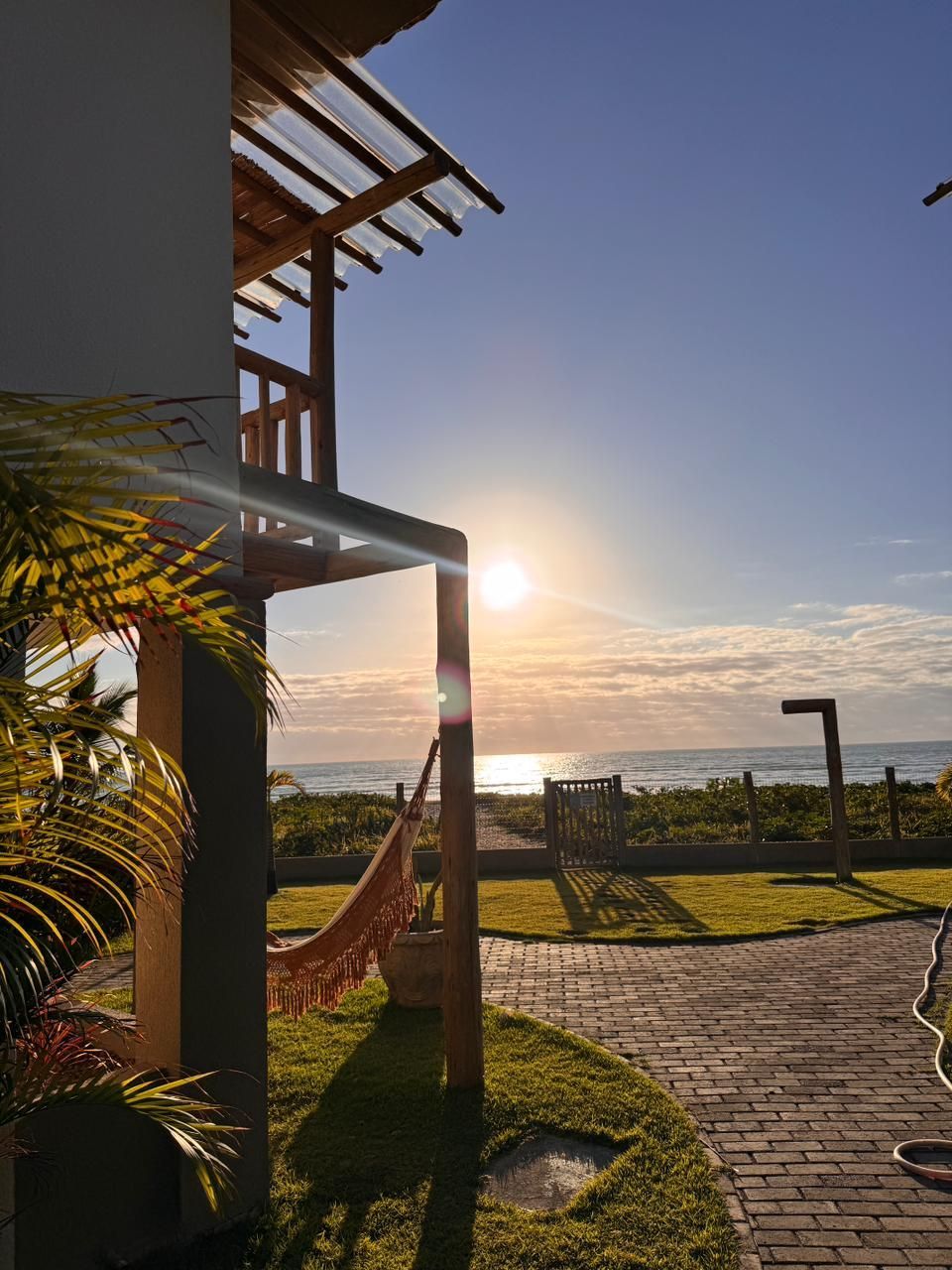 Uma rede está pendurada em uma varanda com vista para uma praia oceânica iluminada pelo sol ao nascer do sol, com um caminho pavimentado na grama nas proximidades.