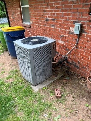 Outdoor HVAC unit beside a brick house wall, near trash bins and an electrical box.