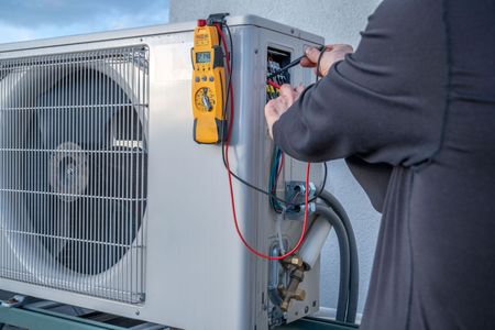 Technician testing wiring on an outdoor HVAC unit with a yellow multimeter