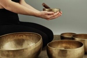 A person sitting and holding a small singing bowl and mallet, with several larger bowls arranged on the floor nearby.
