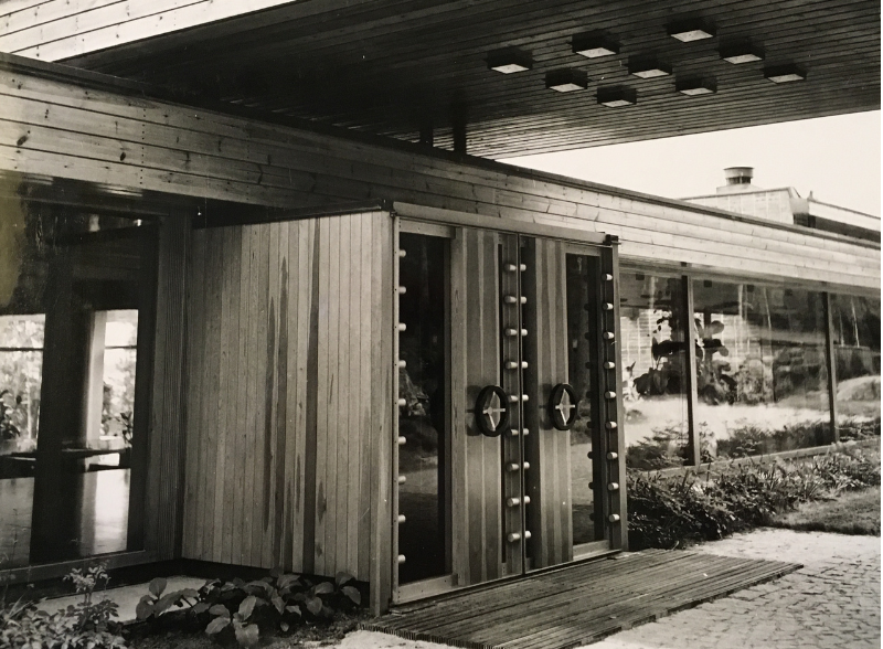 A low-angle, black-and-white photo of a mid-century modern building entrance with wooden paneling and a large overhang.