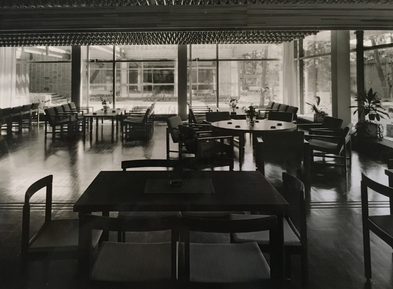 A spacious, dimly lit room with rows of wooden chairs and tables, large floor-to-ceiling windows, and a grid ceiling.