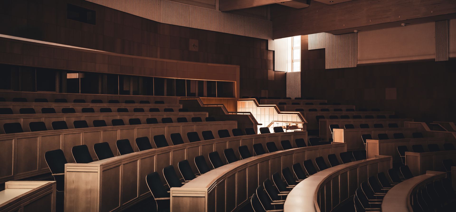 Rows of empty chairs arranged in curved tiers within a dimly lit, wood-paneled lecture hall or assembly room.