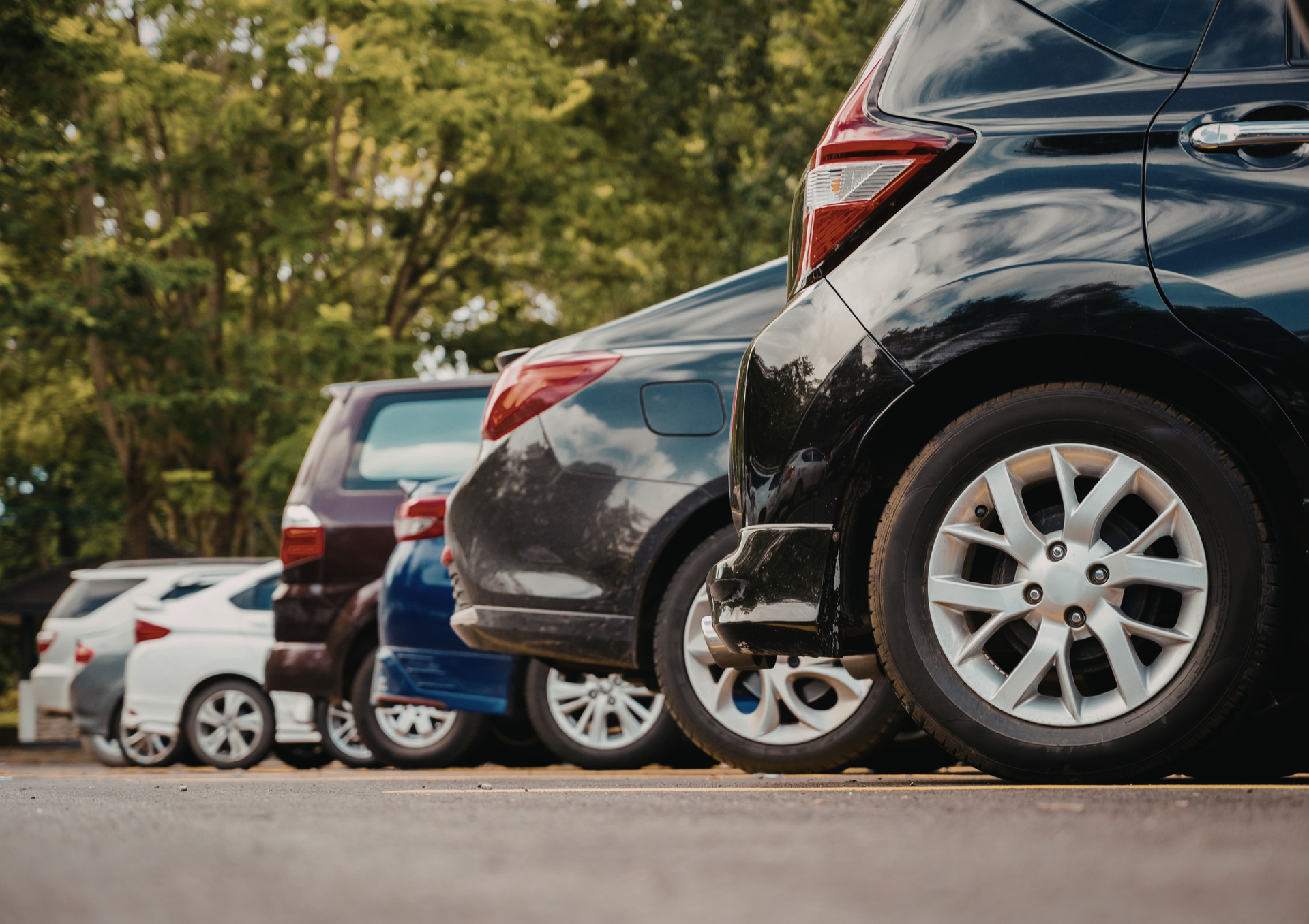 A row of parked cars viewed from behind, featuring a black car in the foreground with alloy wheels in a parking lot.
