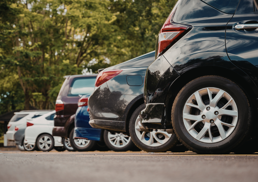 Rivi pysäköityjä autoja takaapäin nähtynä, rivissä pysäköintialueella puita taustalla.