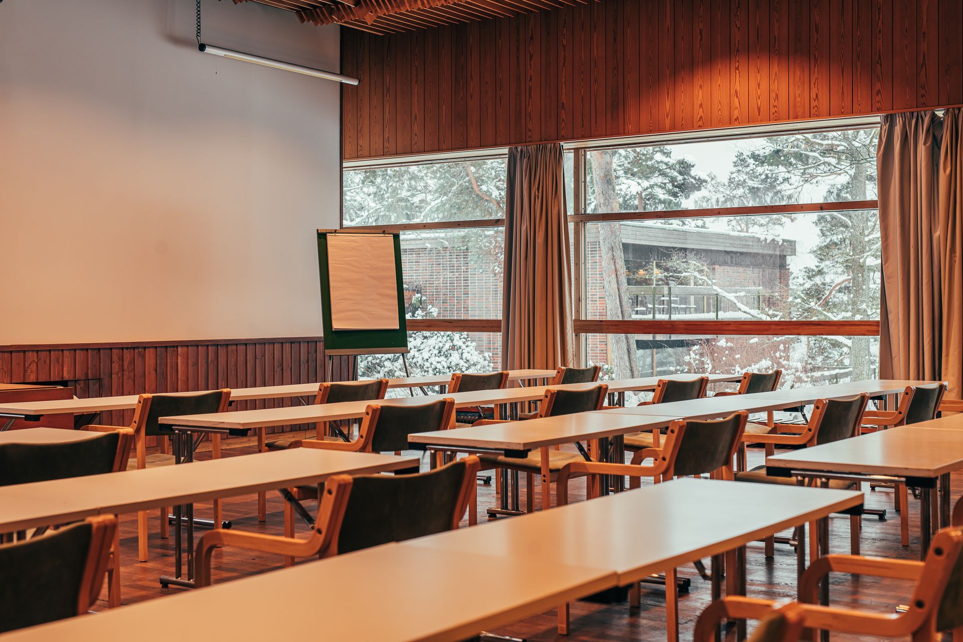 Empty conference room with rows of wooden tables, chairs, and a whiteboard, facing windows overlooking a snowy forest.