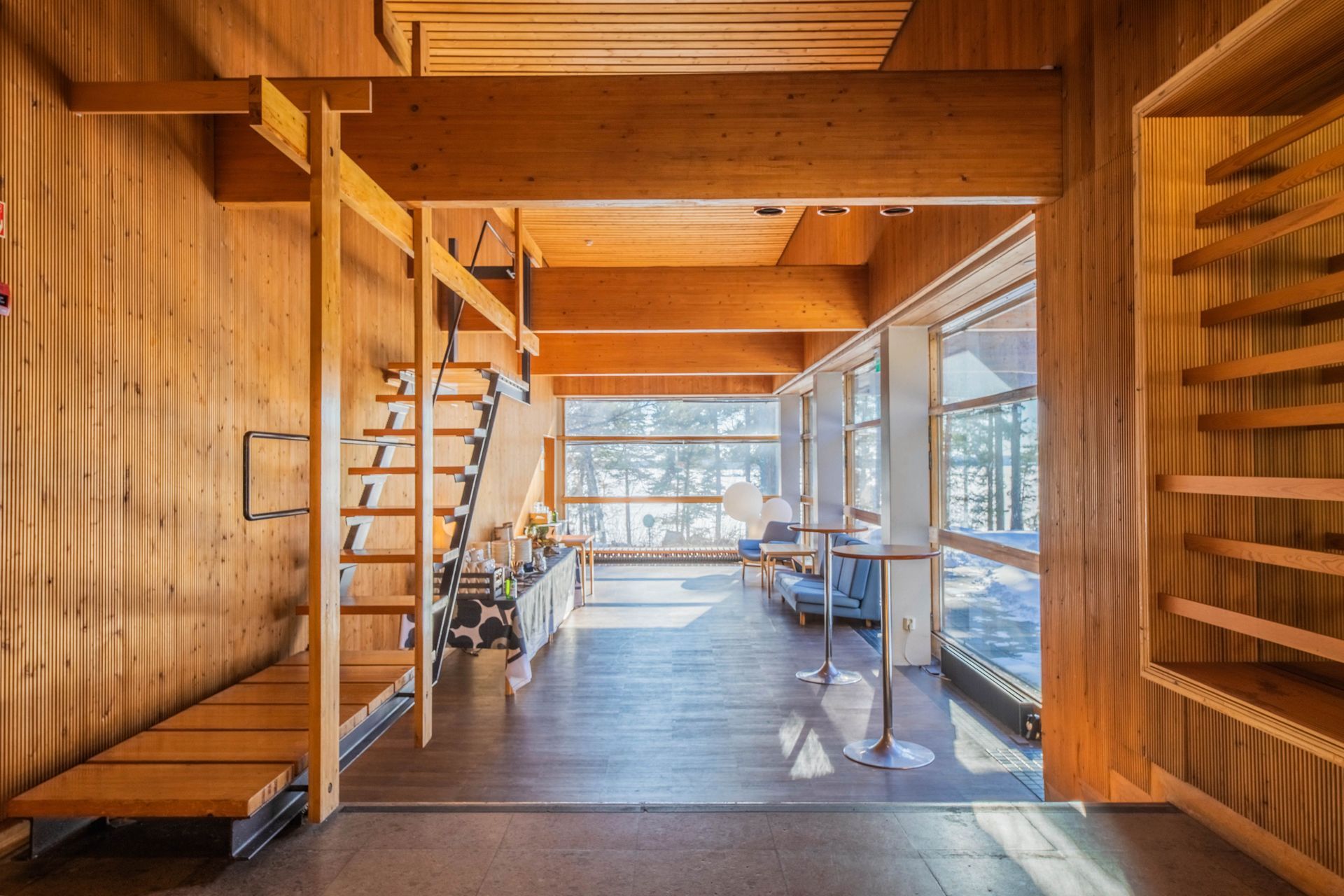 Sunlit indoor area with wooden walls, stairs, and shelving leading to a view of trees through large glass windows.