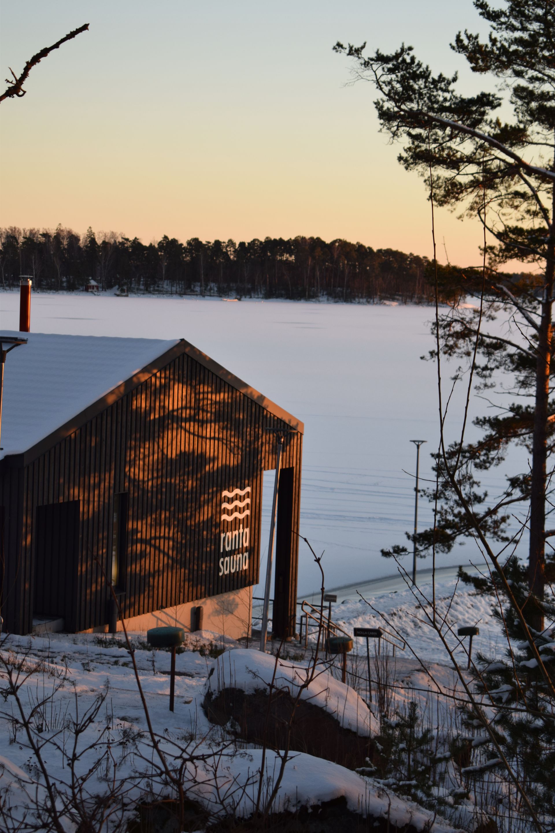 A dark wooden cabin with a logo sits in a snowy landscape by a frozen lake at sunset, flanked by pine trees.