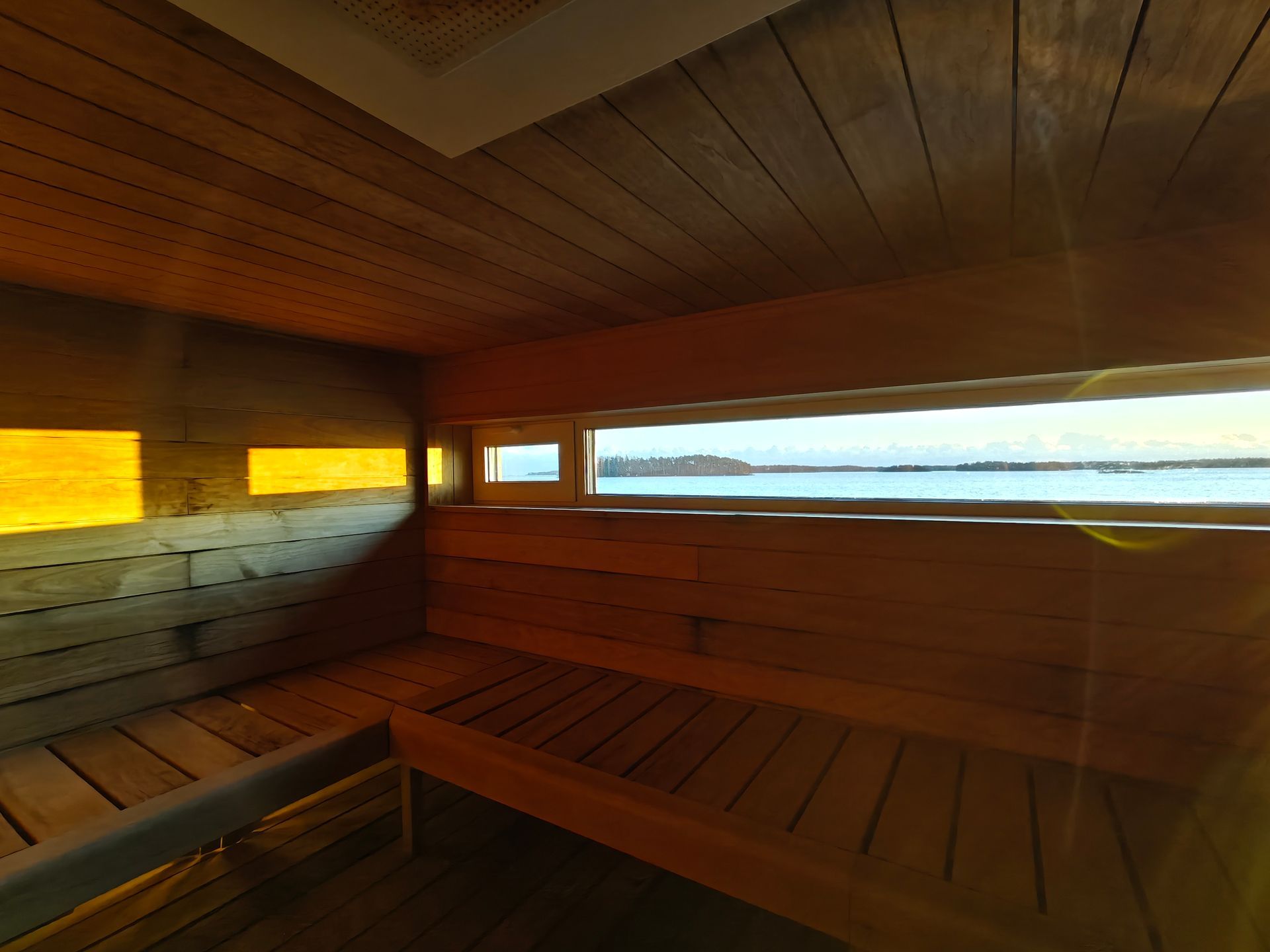 An interior view of a wood-paneled sauna with tiered benches and a wide window looking out toward a body of water.