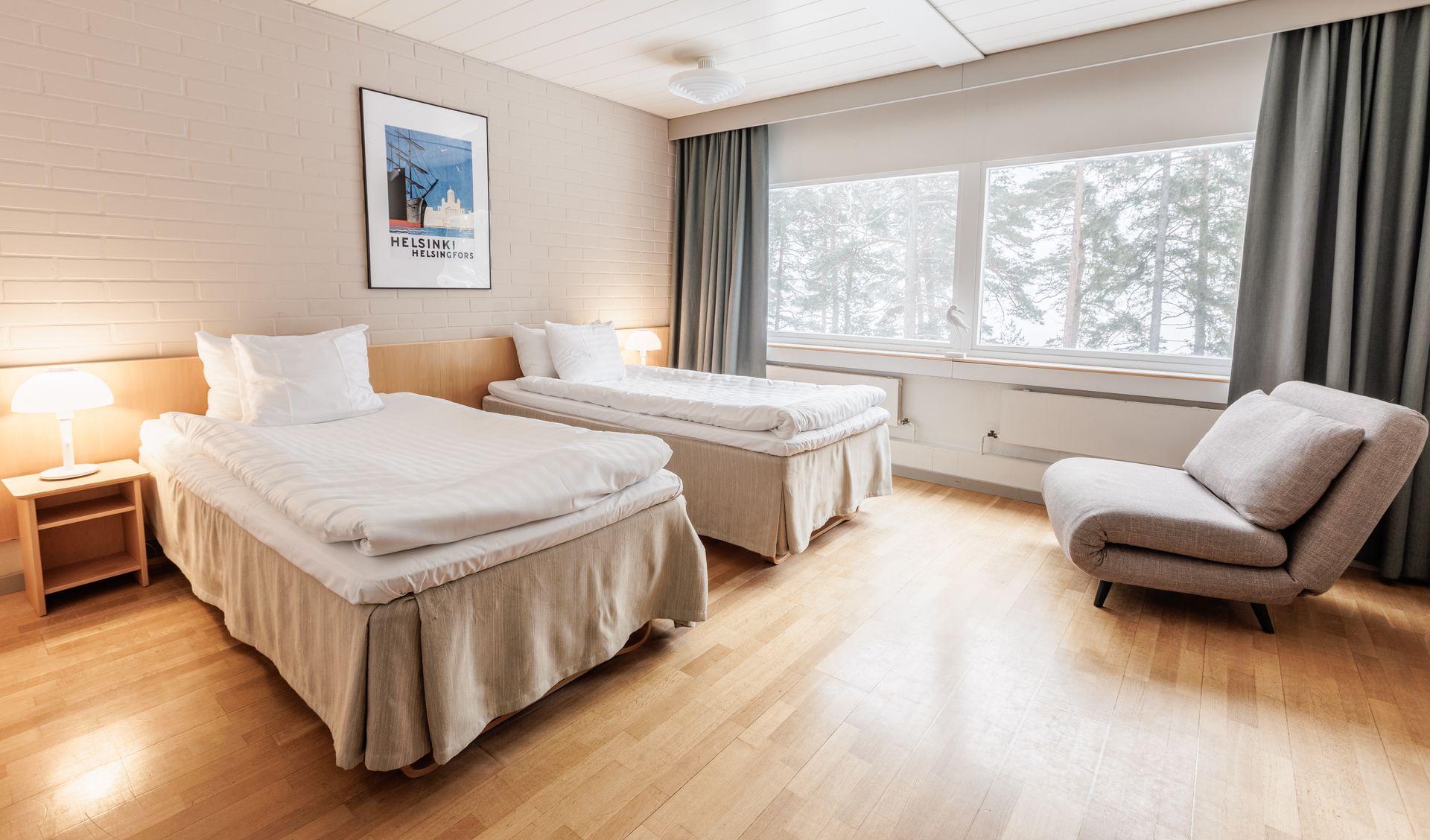 Kirkas hotellihuone, jossa on kaksi erillistä sänkyä, pieni yölamppu ja harmaa nojatuoli suuren ikkunan vieressä, josta on näköala puiden yli.