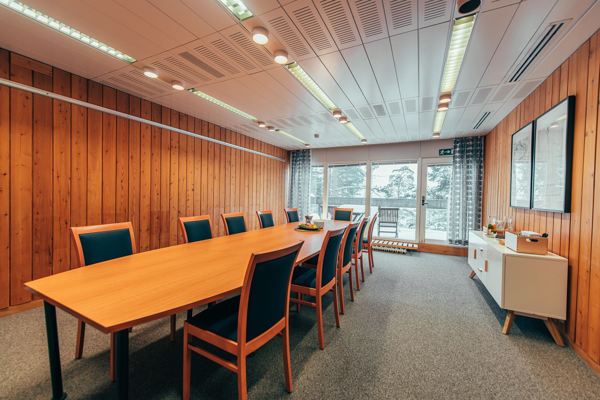 A wood-paneled meeting room with a long table, green chairs, a white sideboard, and large windows with gray curtains.