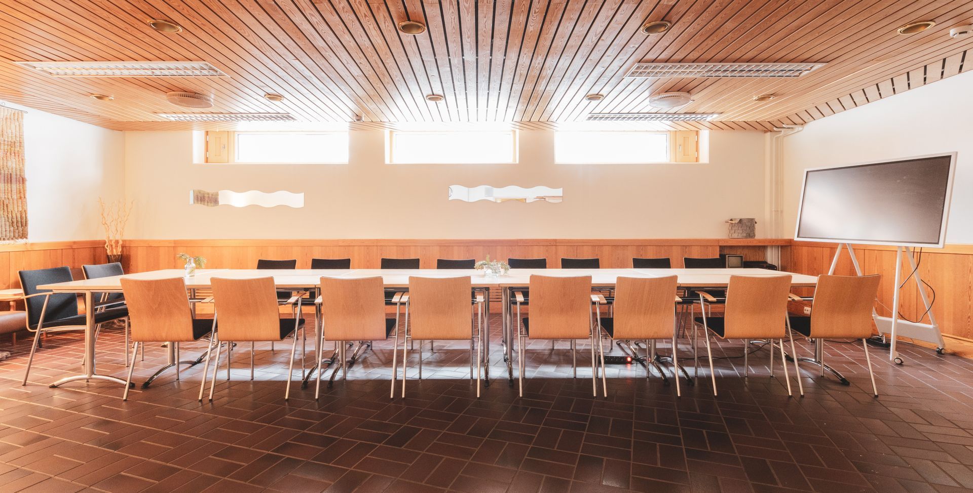 A brightly lit conference room features a long table with ten wooden chairs, a wooden-slatted ceiling, and a flip chart.