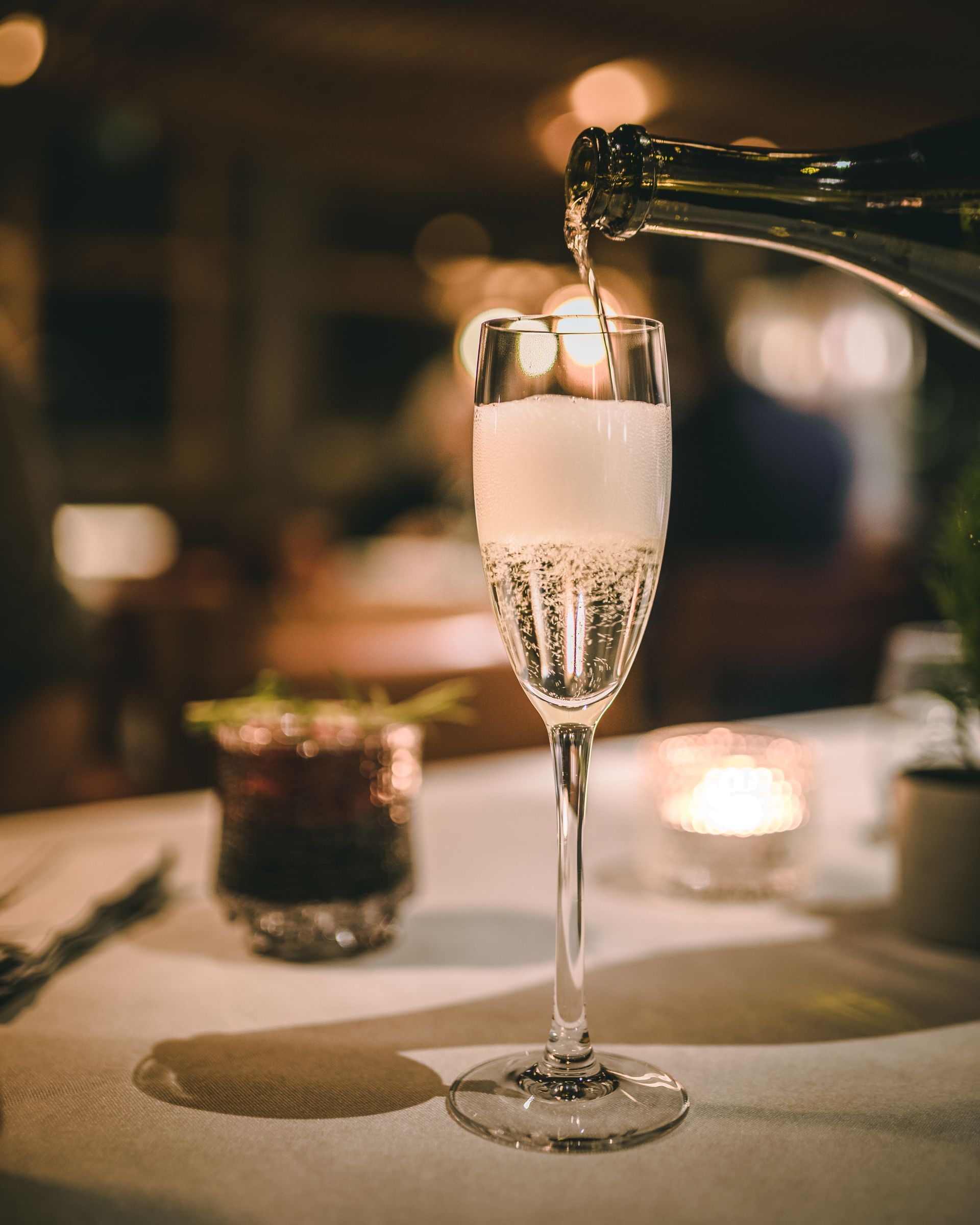 Sparkling wine is poured from a bottle into a tall, elegant glass on a white table in a softly lit, warm setting.