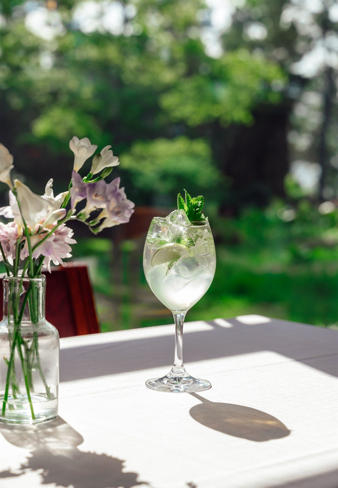 Lasillinen jääcocktailia ja pieni kukkamaljakko ovat pöydällä ulkona luonnonvalossa.