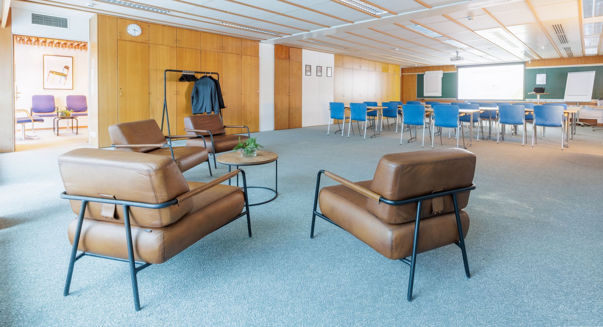 A modern meeting room features two leather armchairs in the foreground and a classroom setup with blue chairs behind.